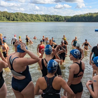 Eine Gruppe von Menschen badet und steht in einem großen, ruhigen See, umgeben von grüner Natur.