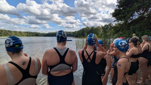 Eine Gruppe von Menschen steht an einem Flussufer mit Blick auf das Wasser und den bewaldeten Uferstreifen.